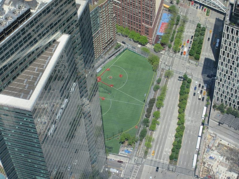 The Battery Park City ball fields seen from a tower high above, a green diamond between apartment blocks and the tree-lined lanes of West Street, a glass skyscraper's corner at left