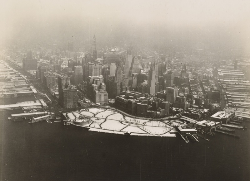 Aerial photograph of the Battery under snow in March 1928, its paths drawn in white, the round Aquarium in Castle Clinton at its western edge, piers on either side and Lower Manhattan's towers in haze behind