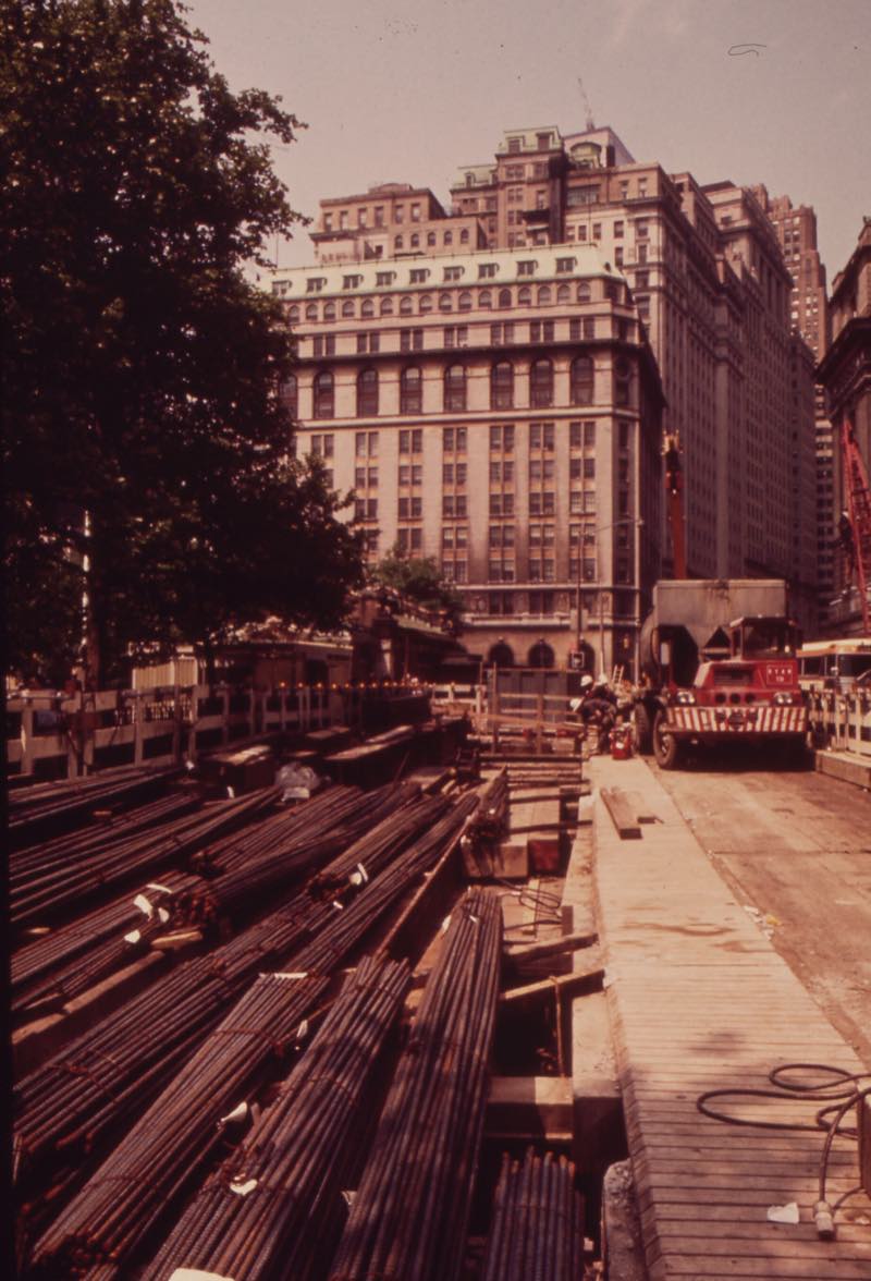 Bundles of steel rebar laid out at a construction site on the east side of the Battery in May 1973, a truck and a crane by the fence and office buildings above