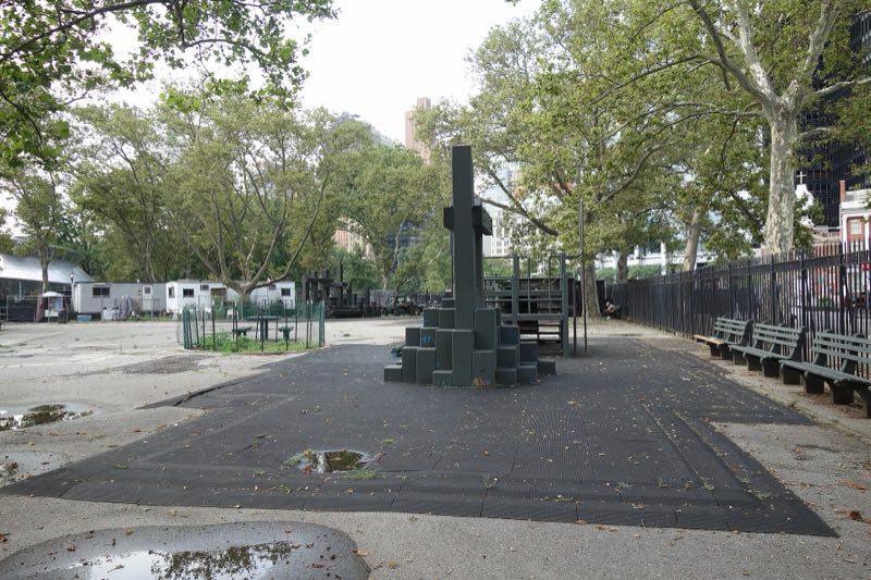 The old Battery playground in July 2019, empty, its rubber surface puddled around a dark climbing structure, benches along an iron fence and construction trailers under the trees behind