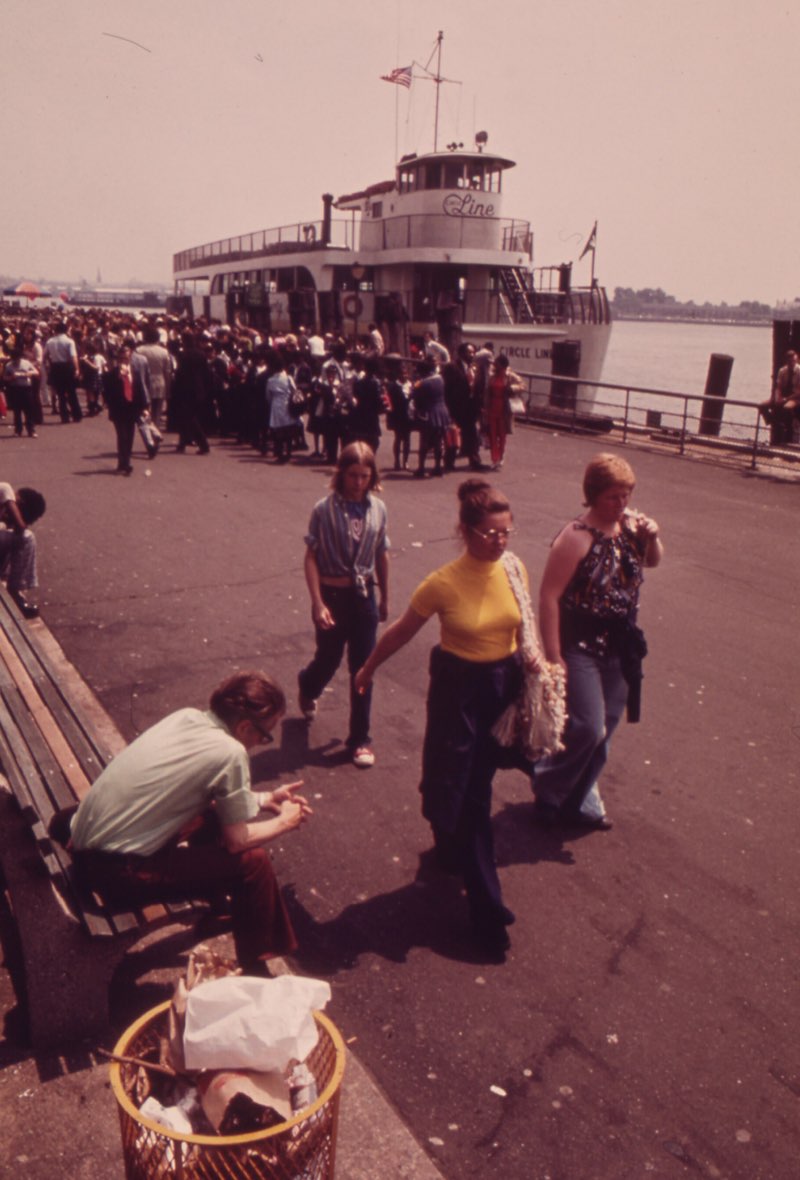 Crowds on the Battery's waterfront in May 1973, a Circle Line boat loading at the pier behind them, a man reading on a bench and three women walking past a wire litter basket