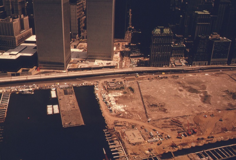 Aerial photograph of the bare Battery Park City landfill in June 1974, a flat expanse of sand and construction vehicles beside the Hudson piers, with the elevated West Side Highway and the new Twin Towers behind