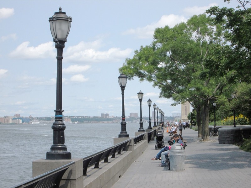 The Battery Park City esplanade on a summer day, cast-iron lamps along the granite river wall, people on benches under the trees and the Hudson at left