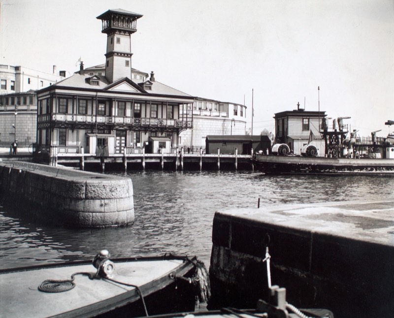 The old fireboat house at the Battery in 1936, a two-story building with a lookout tower on a pier, the boat basin's granite walls and a moored boat in the foreground