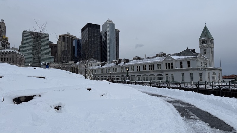 Pier A seen across Wagner Park's snow-covered lawn after the February 2026 blizzard, under a grey sky with Lower Manhattan's towers behind