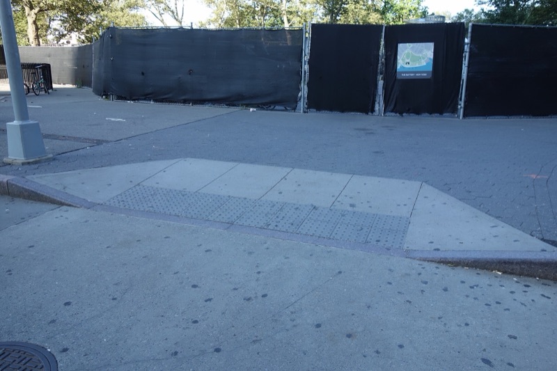 Black-screened construction fence around the playground site at the Battery in August 2019, The Battery's map sign hung on it, an empty sidewalk and curb ramp in front