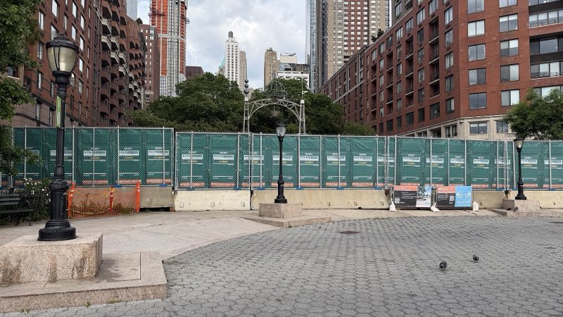 Green-screened construction fence across Rector Place under apartment towers, the BPCA's information and detour boards at its foot, lampposts and pigeons on the empty plaza in front