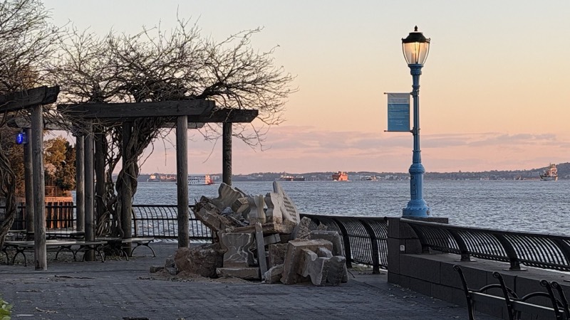 Broken granite blocks stacked on the esplanade beside the South Cove pergola at dusk, a blue lamppost with a banner and the harbor beyond