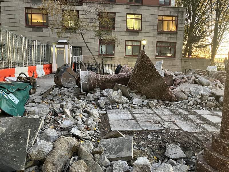 A toppled pebbled column and its pyramid capital lying in rubble where the Upper Room stood, at dusk, apartment windows lit behind