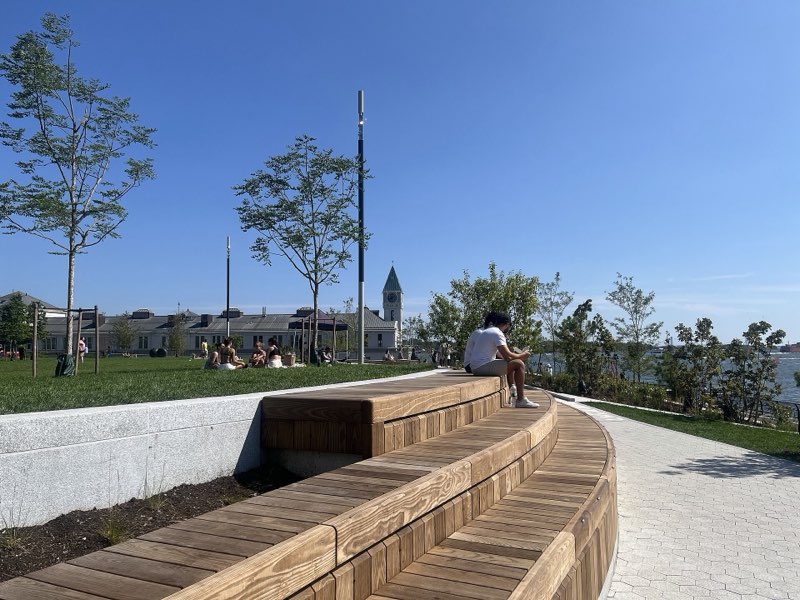 Curved timber benches stepping down from Wagner Park's raised lawn beside its granite edge wall, people on the grass and Pier A's clock tower beyond under a clear sky