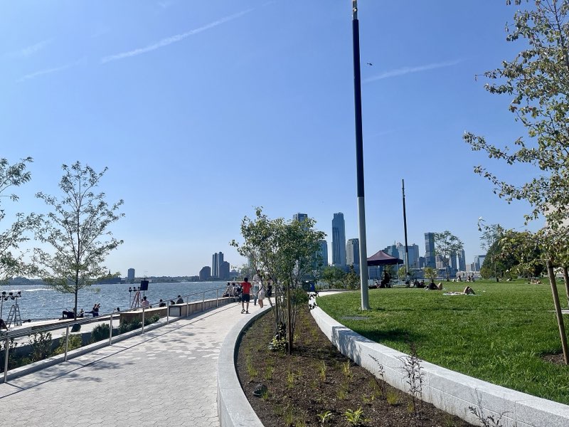 The rebuilt Wagner Park's waterfront path on a clear day, young trees in a new planting bed and a granite curb between the path and the lawn, people on the grass and the harbor at left