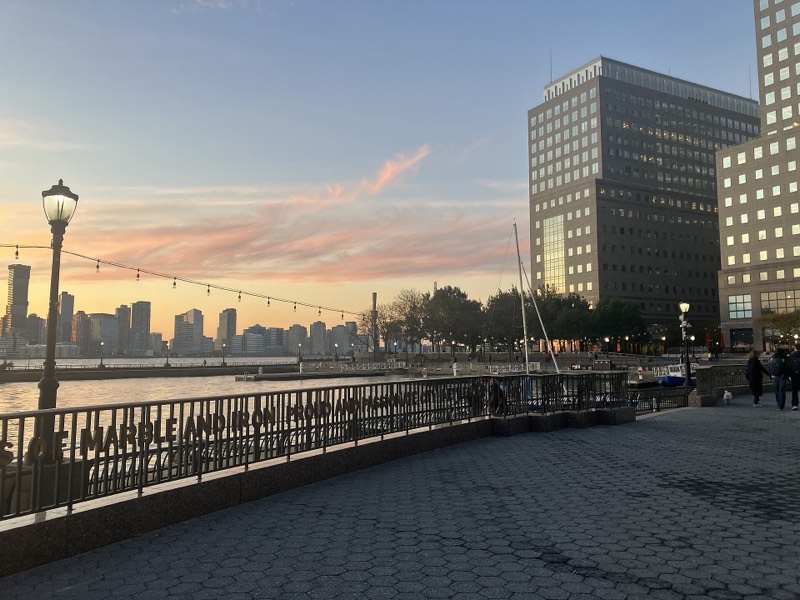 The Waterfront Plaza at North Cove at sunset, string lights over a railing lettered with a quotation, the Hudson and Jersey City at left and office towers with lit windows at right