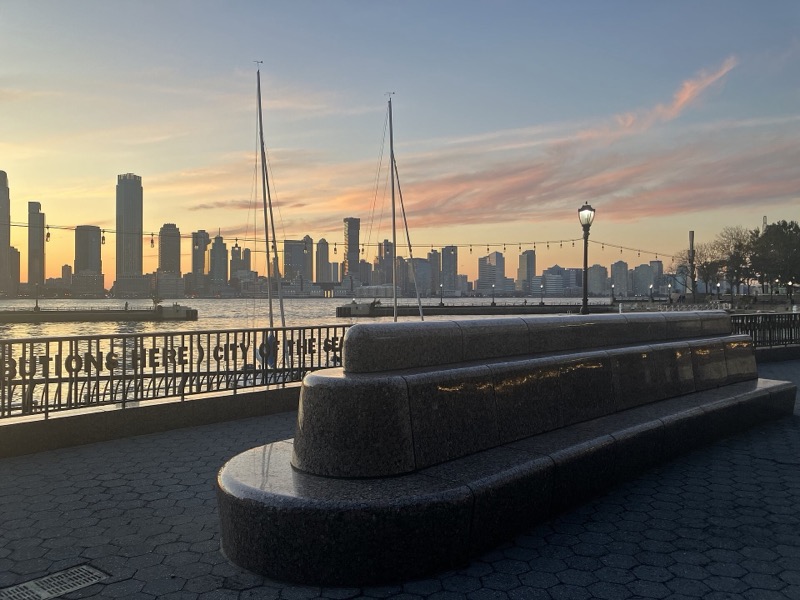 A long polished granite bench on the Waterfront Plaza at dusk, two sailboat masts and the Jersey City skyline across the river under a pink sky